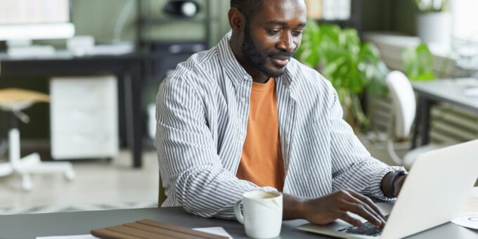 A smiling man in a striped shirt works on a laptop at an office desk, preparing his company's statement of capital online.