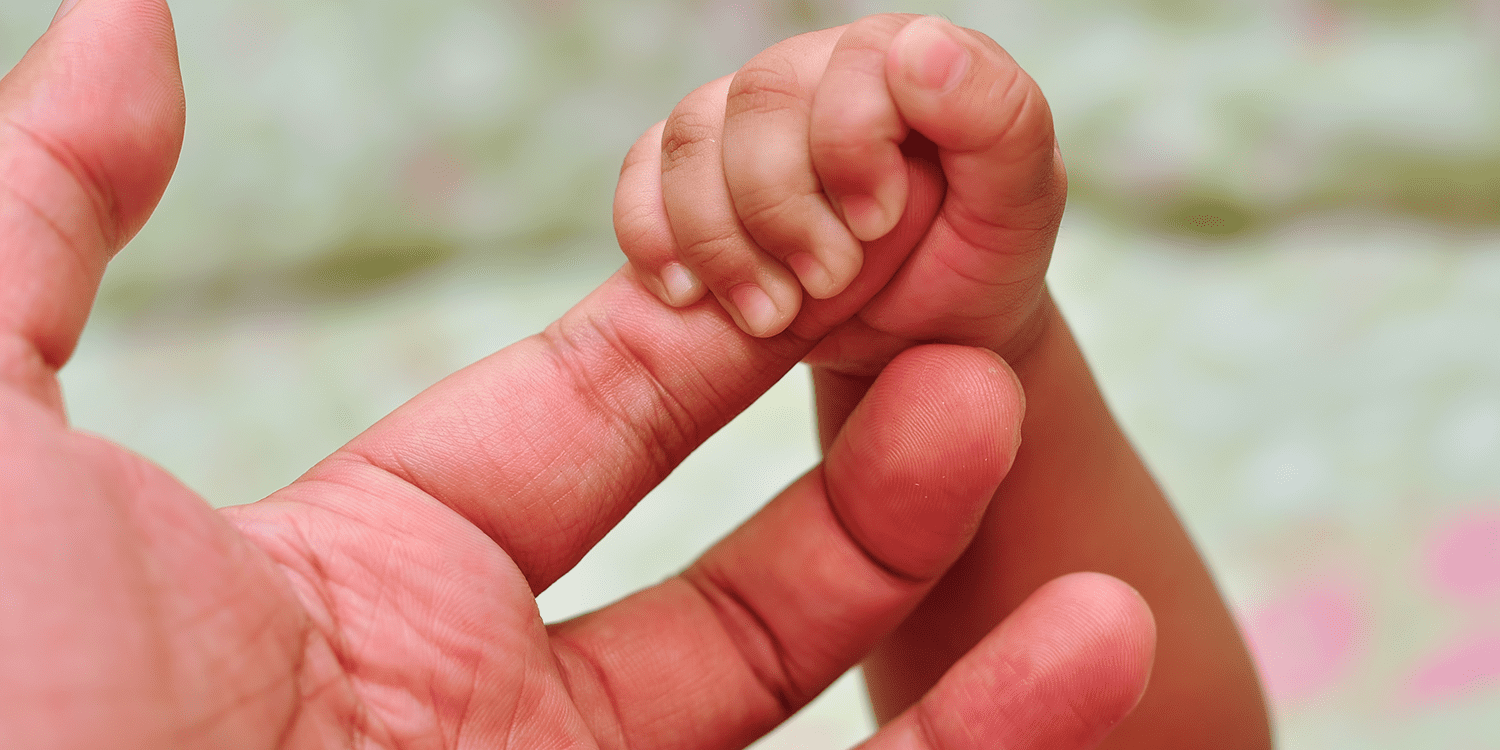 A baby's hand holding holding the finger of an adult's hand, representing the topic of maternity pay.