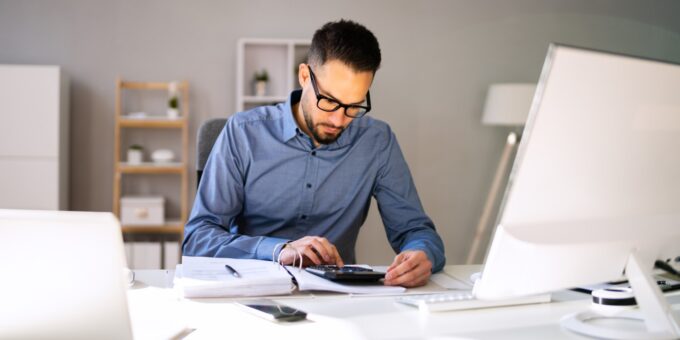 Company director working out how to take money out of a limited company using a calculator and paperwork at his desk.