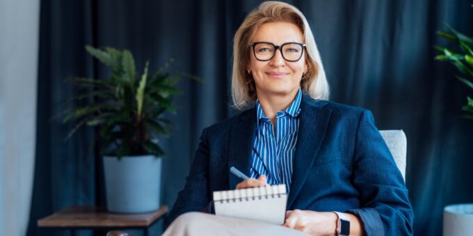 Confident middle-aged woman with blonde hair and glasses sitting in a modern office space, smiling at the camera while holding a pen and notebook. She is wearing a navy blazer over a blue-striped shirt and an Apple Watch. Two potted plants and a curtain are visible in the background. She is a subscriber of a company.