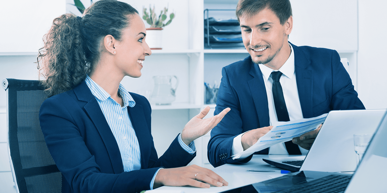 Image of a man and a woman in business attire, sitting at a desk and discussing work, representing the collaborative nature of the relationship between a company director and a company secretary.