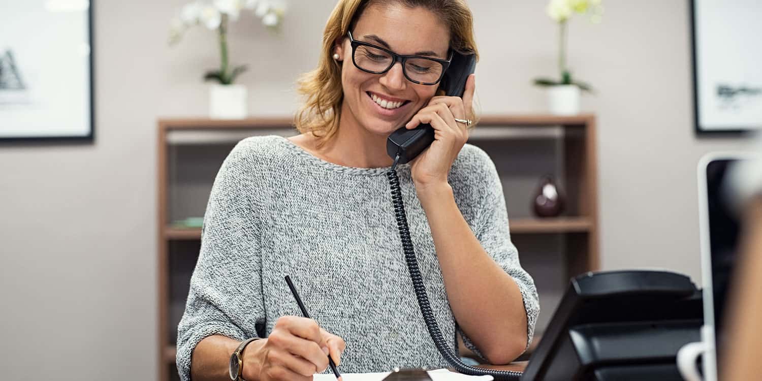 Image of a woman holding a telephone handset to her ear whilst sitting at her desk, illustrating the concept of a business landline telephone number.