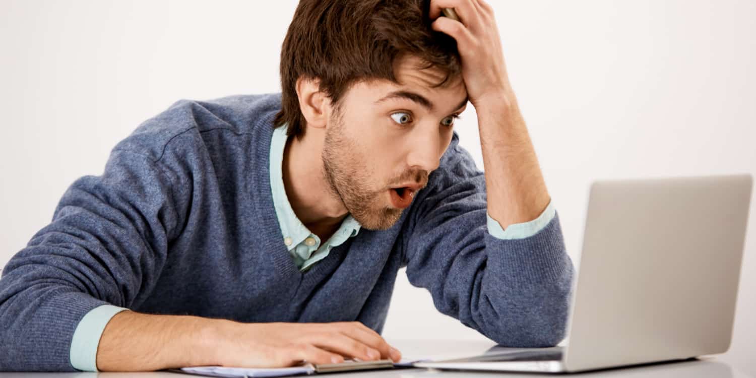 Young businessman looking at his laptop screen horrified and surprised by mistakes he has made.