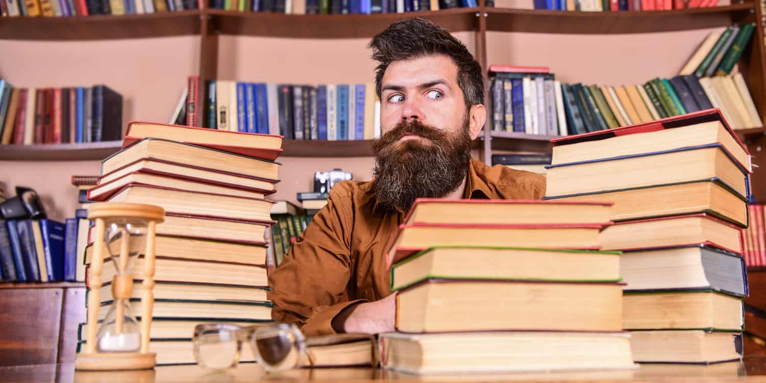 Young man with beard, sitting at a desk and surrounded by stacks of books.