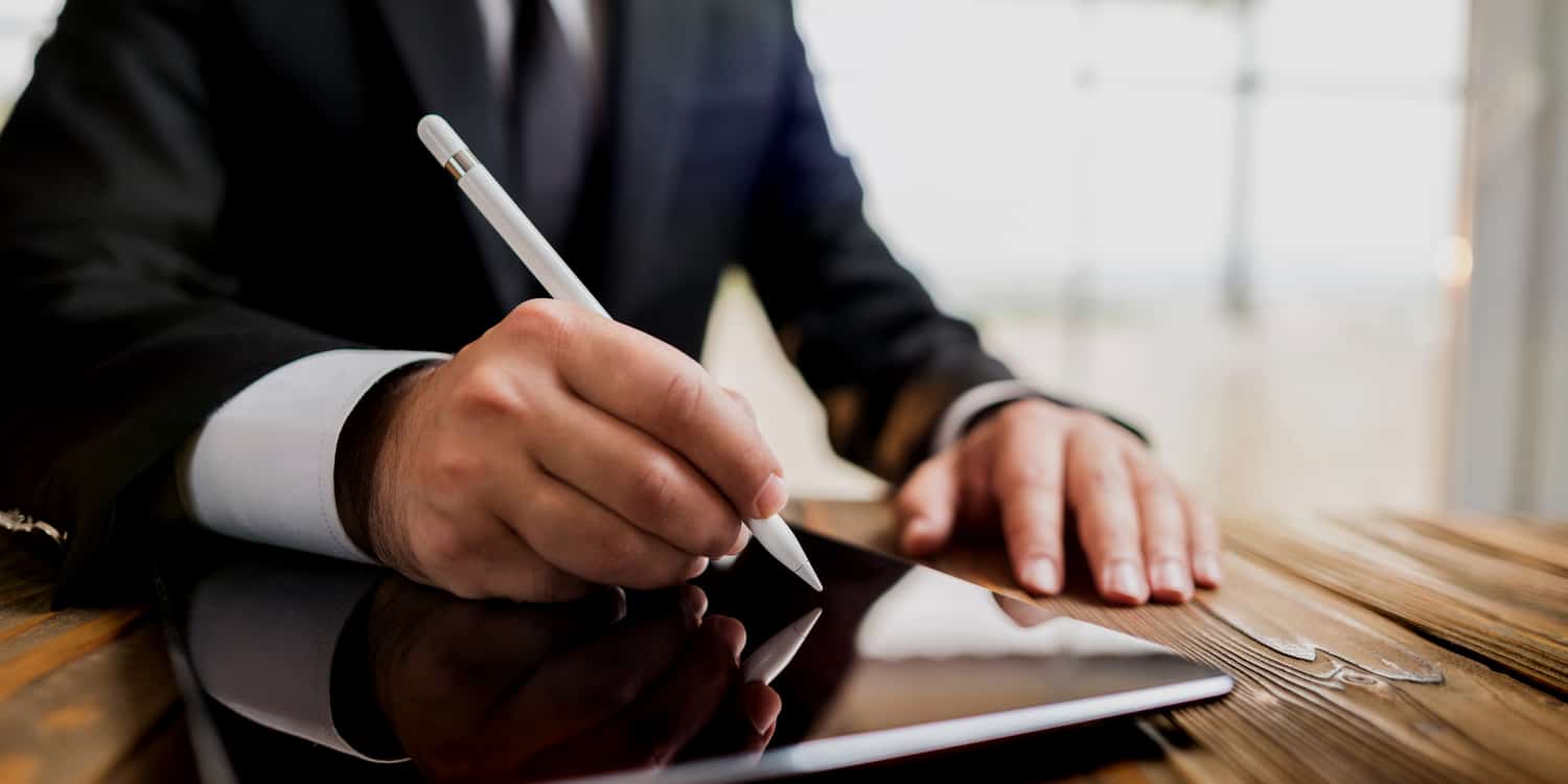 Businessman sitting at desk signing a document on an iPad using a light pen - illustrating the electronic signature concept.