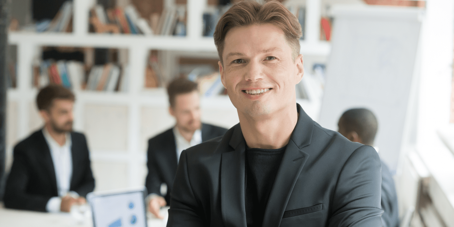 Young male director smiling at camera with colleagues in background.