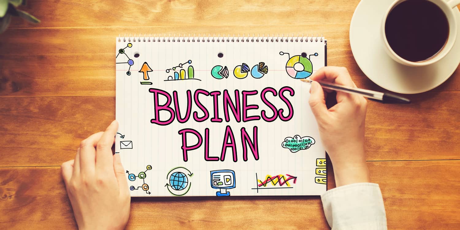 Business Plan text written on a pad of paper with a person holding a pen on a wooden desk.