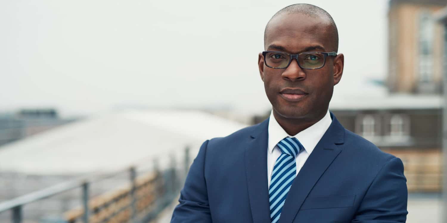 Confident sole director in a stylish suit standing with folded arms on a rooftop of an office block looking at the camera with a serious expression.