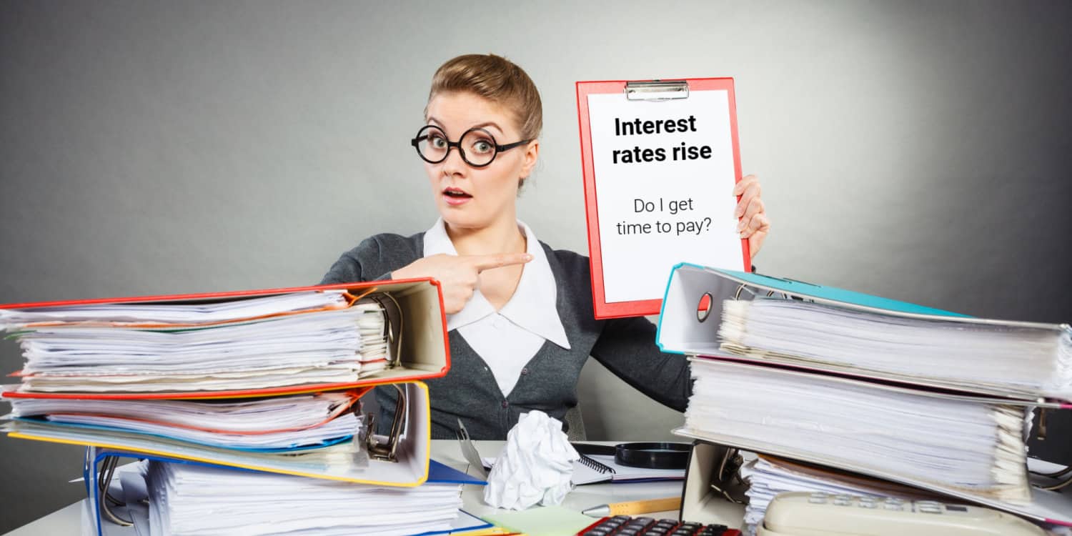 Businesswoman sitting at a desk behind files, holding up and pointing to a clipboard with headline 'Interest rates rise' and sub-heading of 'Do I get time to pay?'