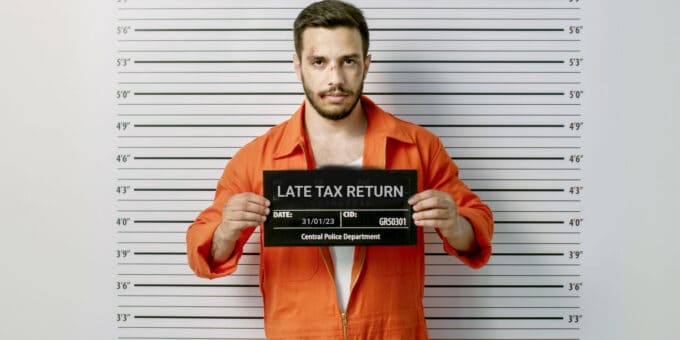 Man in a police station wearing prisoner orange jumpsuit and holding placard with headline LATE TAX RETURN. Height Chart in the background.