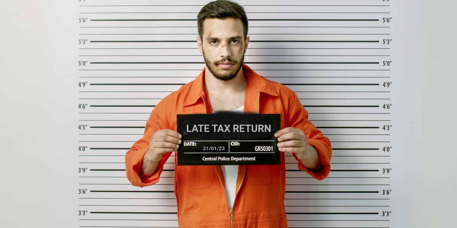 Man in a police station wearing prisoner orange jumpsuit and holding placard with headline LATE TAX RETURN. Height Chart in the background.