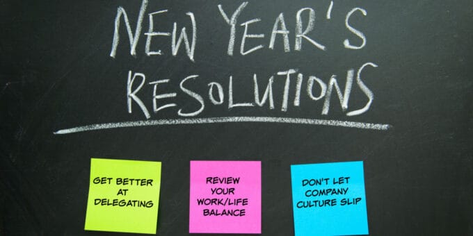 The word New Year's Resolutions written on the blackboard with 3 postit notes displaying business resolution ideas.
