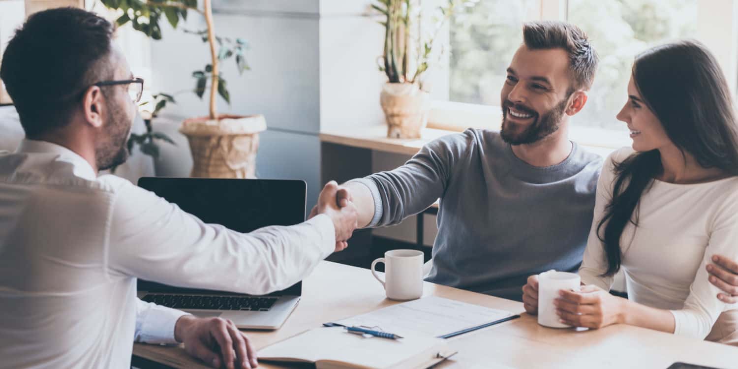 A young businessman sitting next to his wife and shaking hands with a man sitting opposite him at a desk. Illustrating the concept of sealing a deal in selling his business.