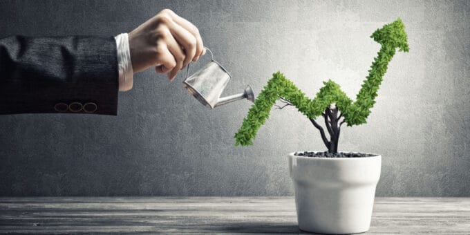 Hand of a businessman watering a small plant in a pot shaped like a growing graph.