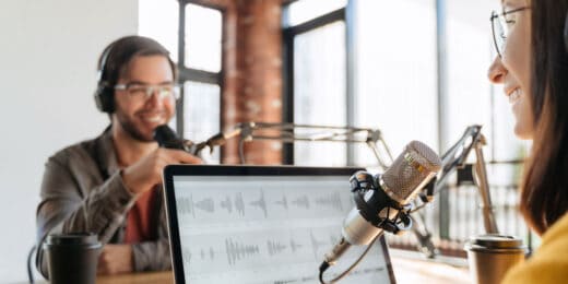A man and woman conducting a podcast in headphones looking at each other while talking into large microphones.
