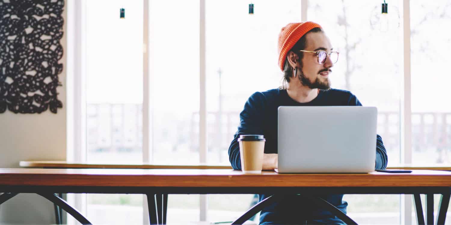 Young trendy male looking away and thinking about starting his own freelance copywriting business, while on laptop computer in coffee shop.