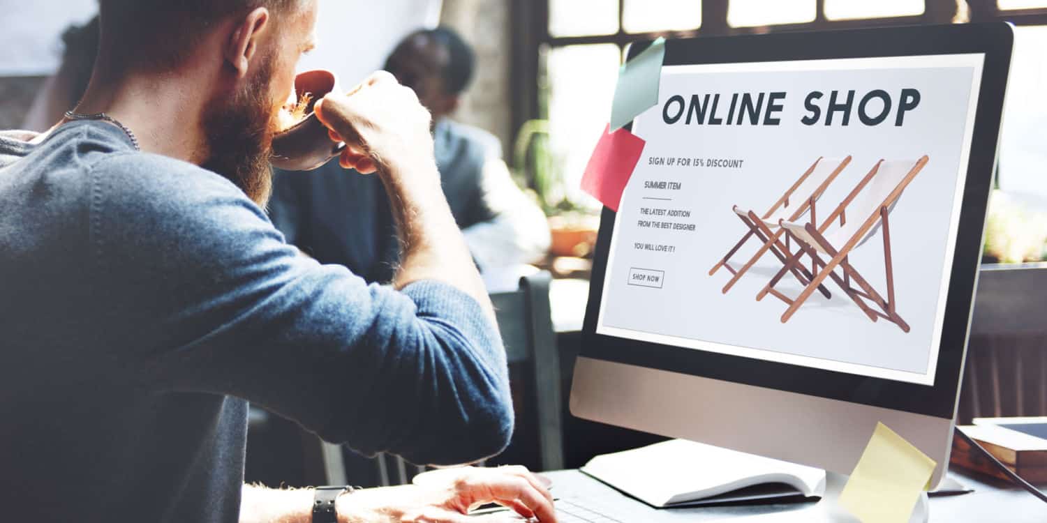 Young male with beard drinking coffee and browsing an online shop at his office desk.