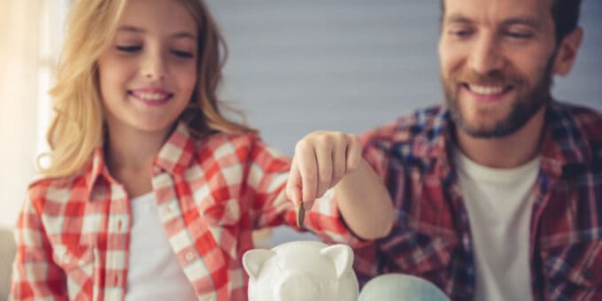 Pretty girl is putting a coin into a piggy bank that her father is holding. Concept of family investment.