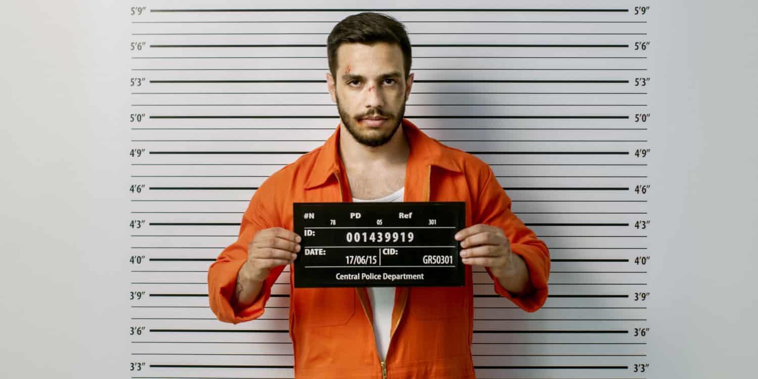 Man getting front-view mug shot in a police station, wearing orange jump suit, holding placard and with height chart in the background.