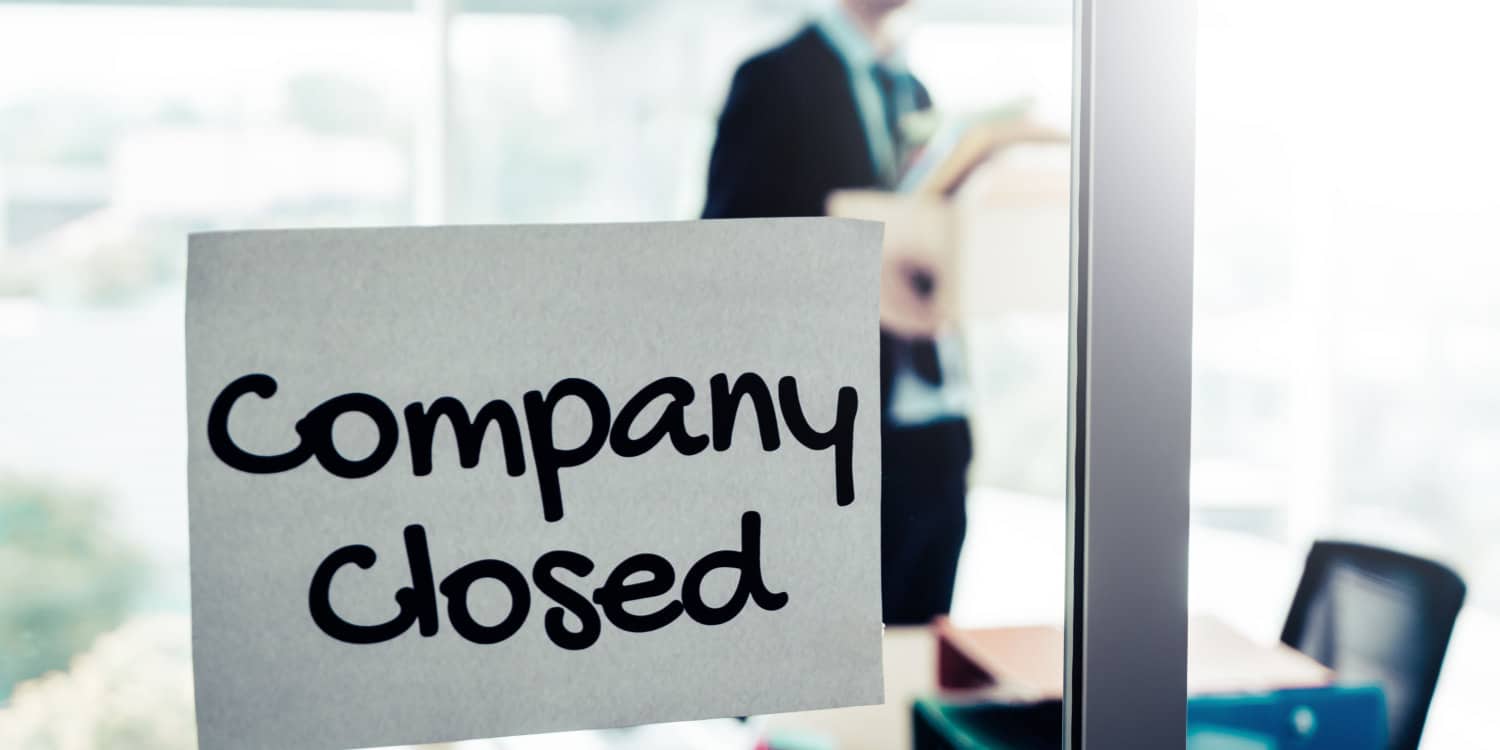 A paper 'Company Closed' sign stuck to the glass window of a meeting room, with a businessman standing in the room