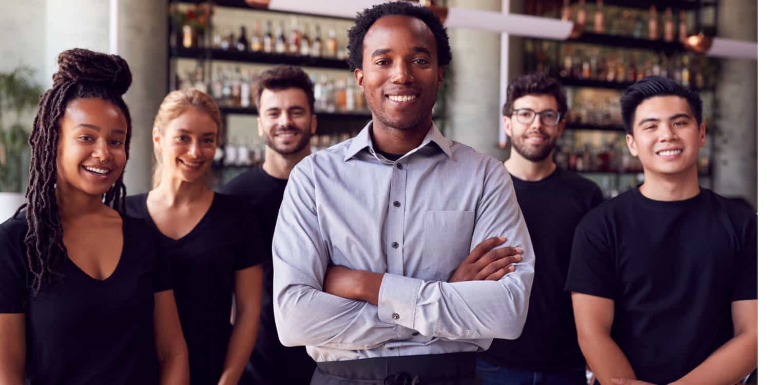 Portrait of a male franchise owner standing in his restaurant with his team of waiting staff standing behind him.