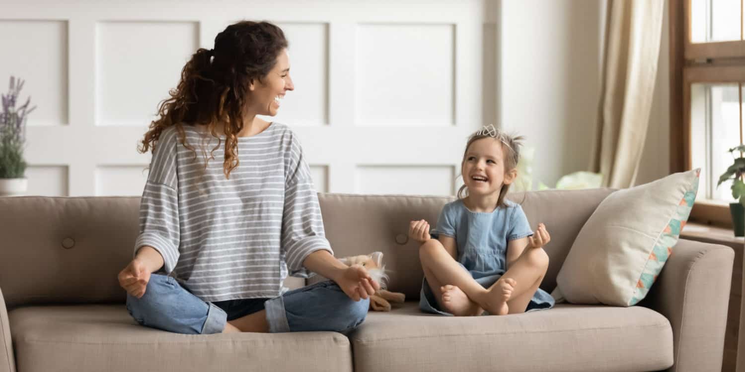 Smiling female child minder sitting on comfortable couch with cute playful little girl in lotus position.