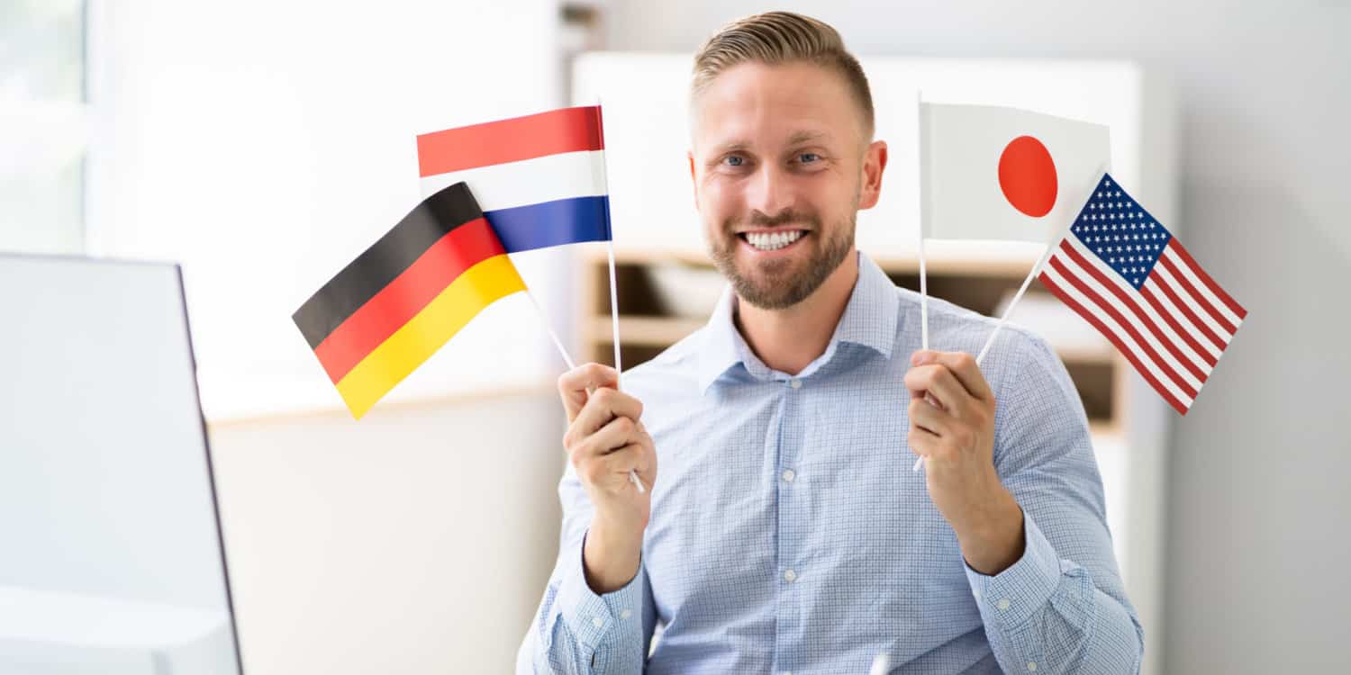 Young businessman sitting in his office at a desk, and holding up 4 foreign flags.