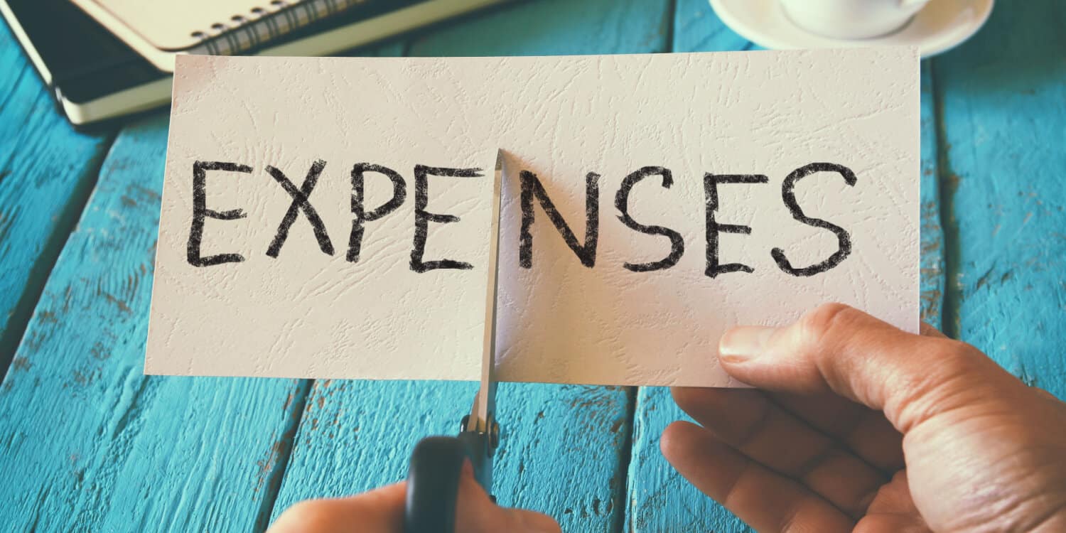A man hand holding a card with the word EXPENSES written in black capital letters, and cuting the card in half with a pair of scissors.