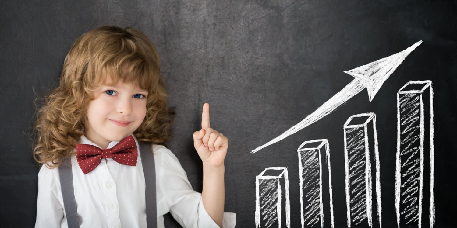 Smiling young girl wearing a white shirt and bow tie, pointing upwards infront of a blackboard with a bar chart and upwards arrow. Concept of scaling a business.