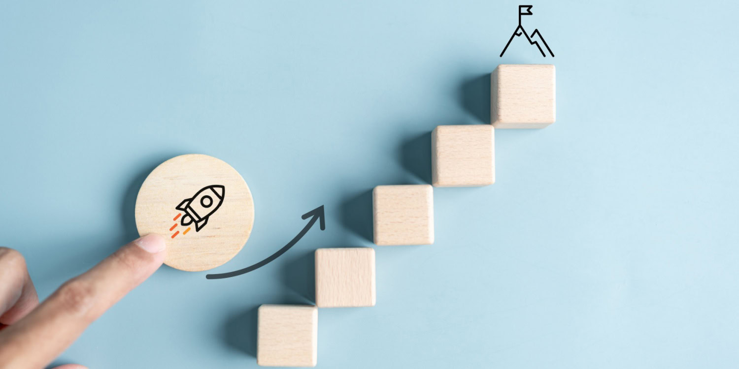 A hand points at a wooden disc with a rocket graphic, showcasing progress toward a mountain peak represented by wooden blocks, symbolising the stages of scaling a business.
