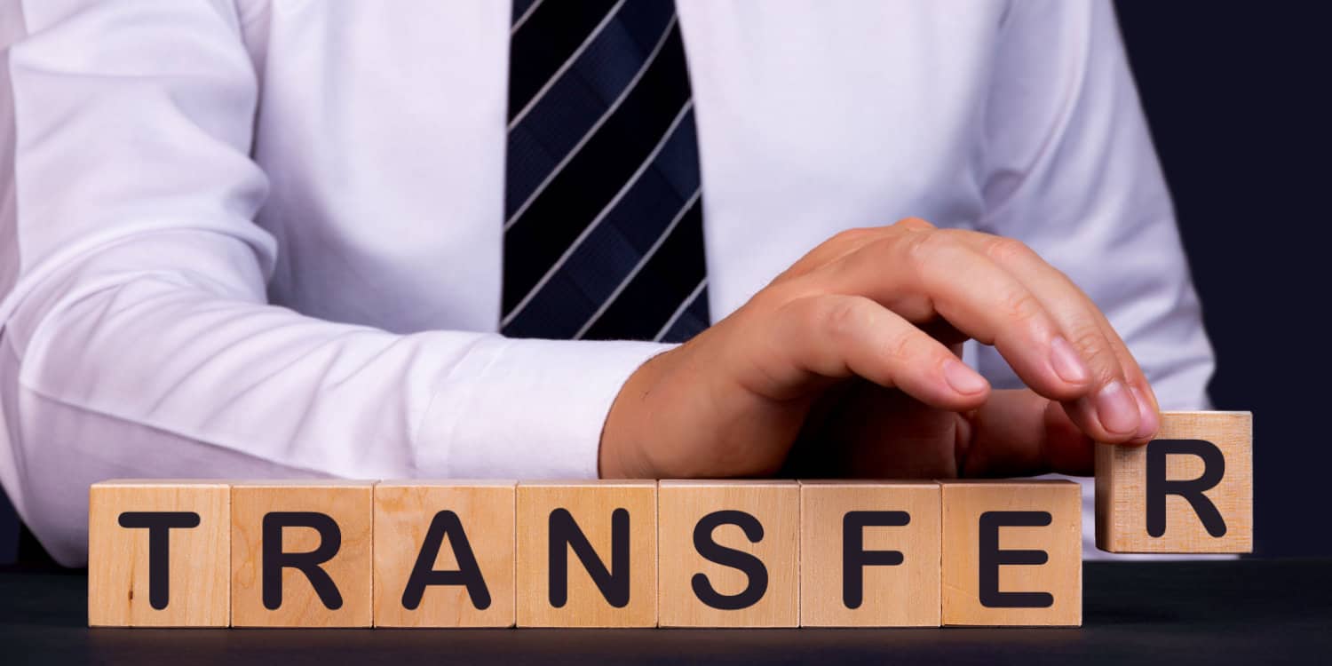 Businessman in white shirt and dark tie laying out wooden blocks on a desk to spell the word TRANSFER.
