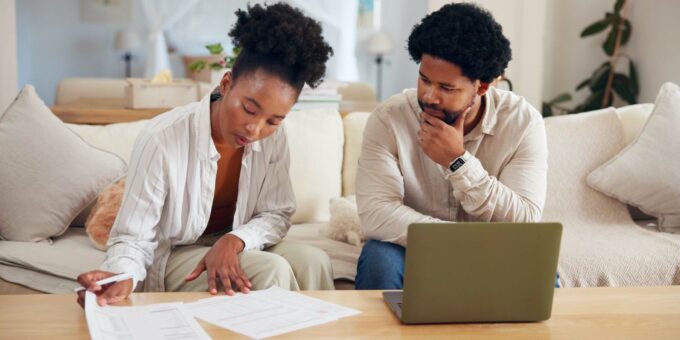A woman reviews paperwork while a man observes thoughtfully, both seated at a table with a laptop in a cozy living room environment