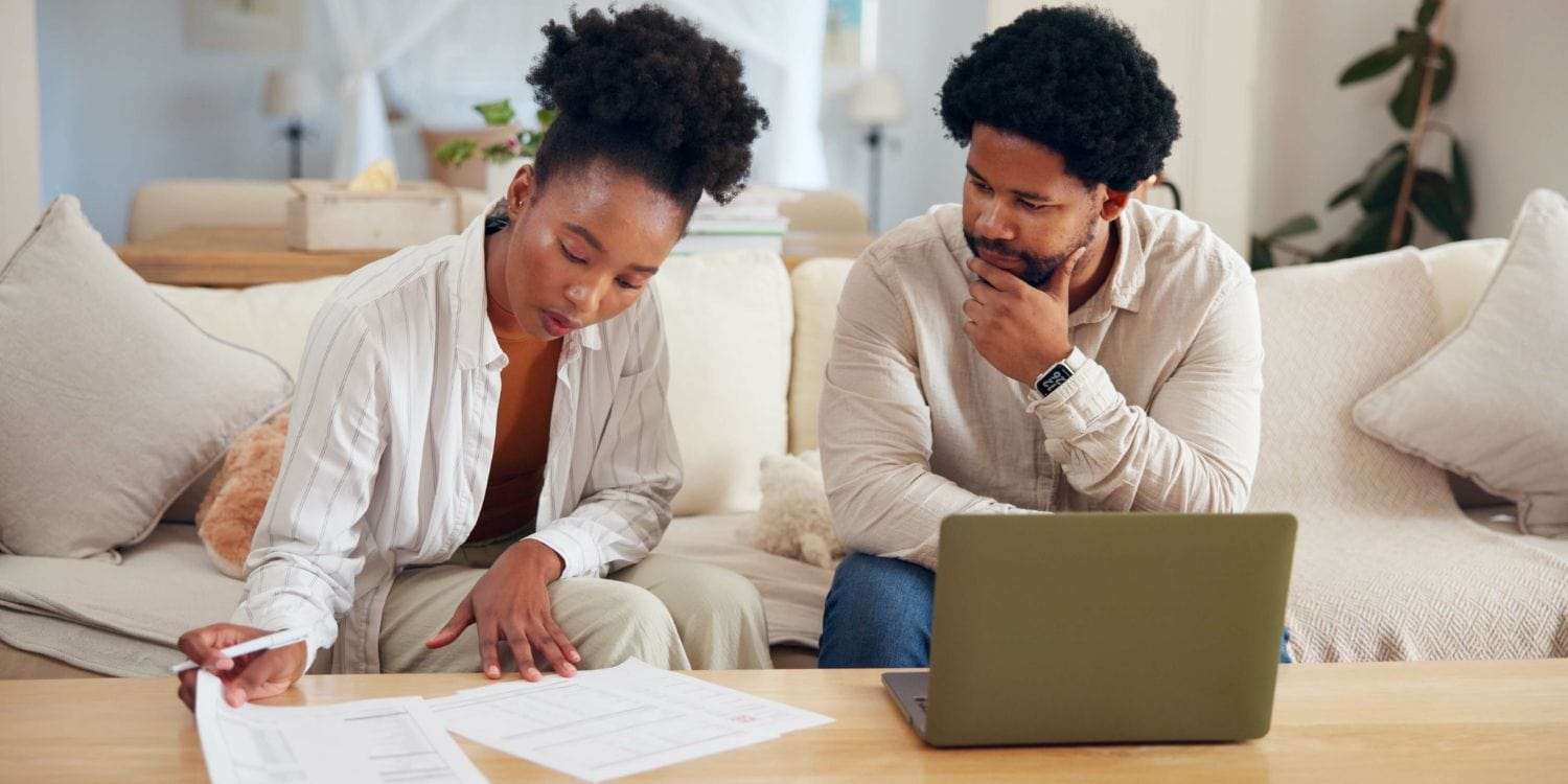 A woman reviews paperwork while a man observes thoughtfully, both seated at a table with a laptop in a cozy living room environment