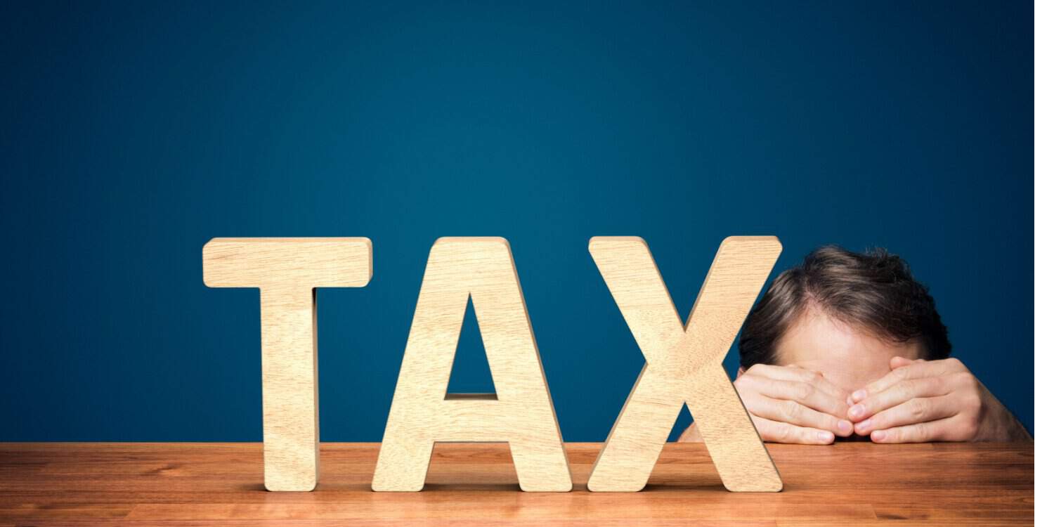 Businessman has a fear of the outcome of his tax returns and is hiding behind a desk with his hands over his eyes. The desk has a large 3d model of the word TAX sitting on it.