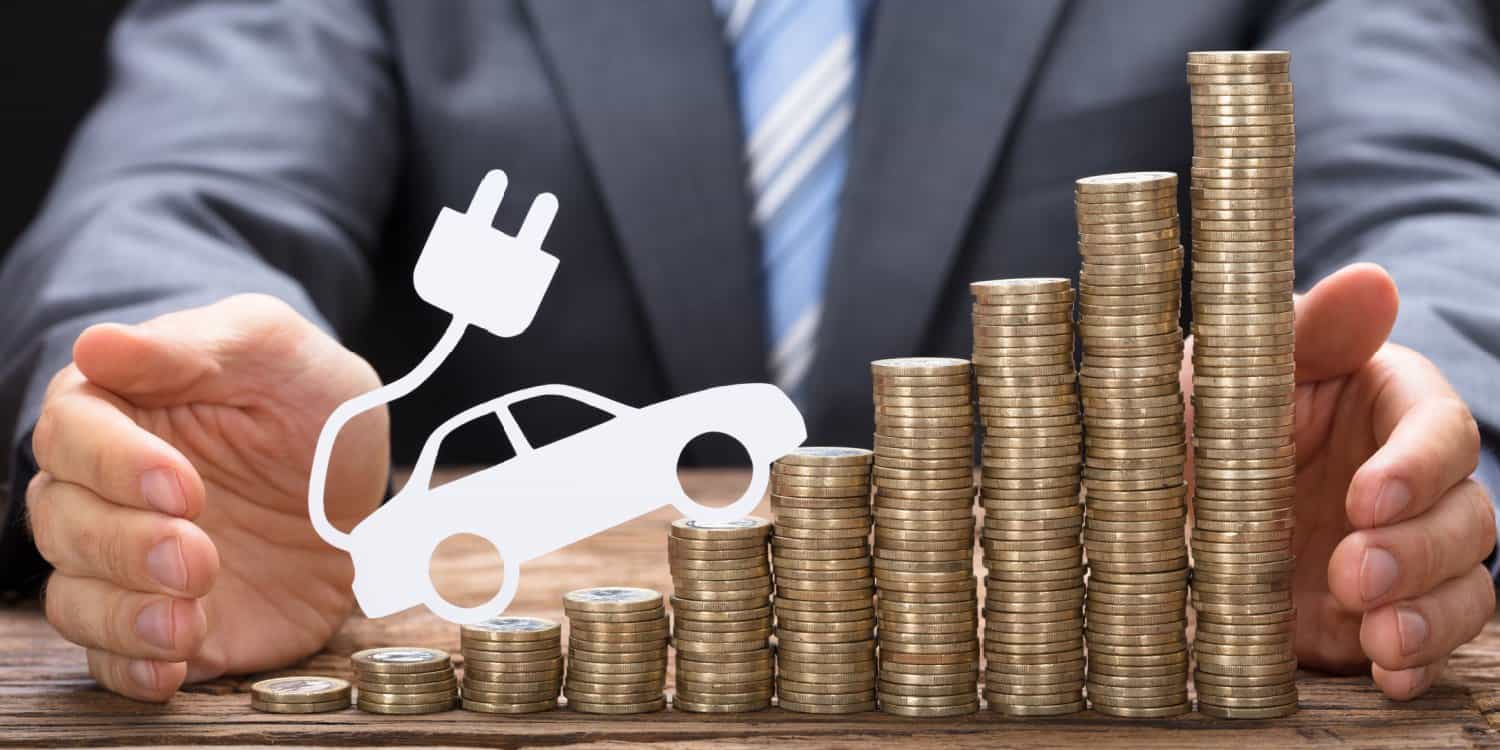 Businessman sitting at a desk with a row of stacked coins rising in height from left to right with an electric car image sitting on top of the coins in an upward position.