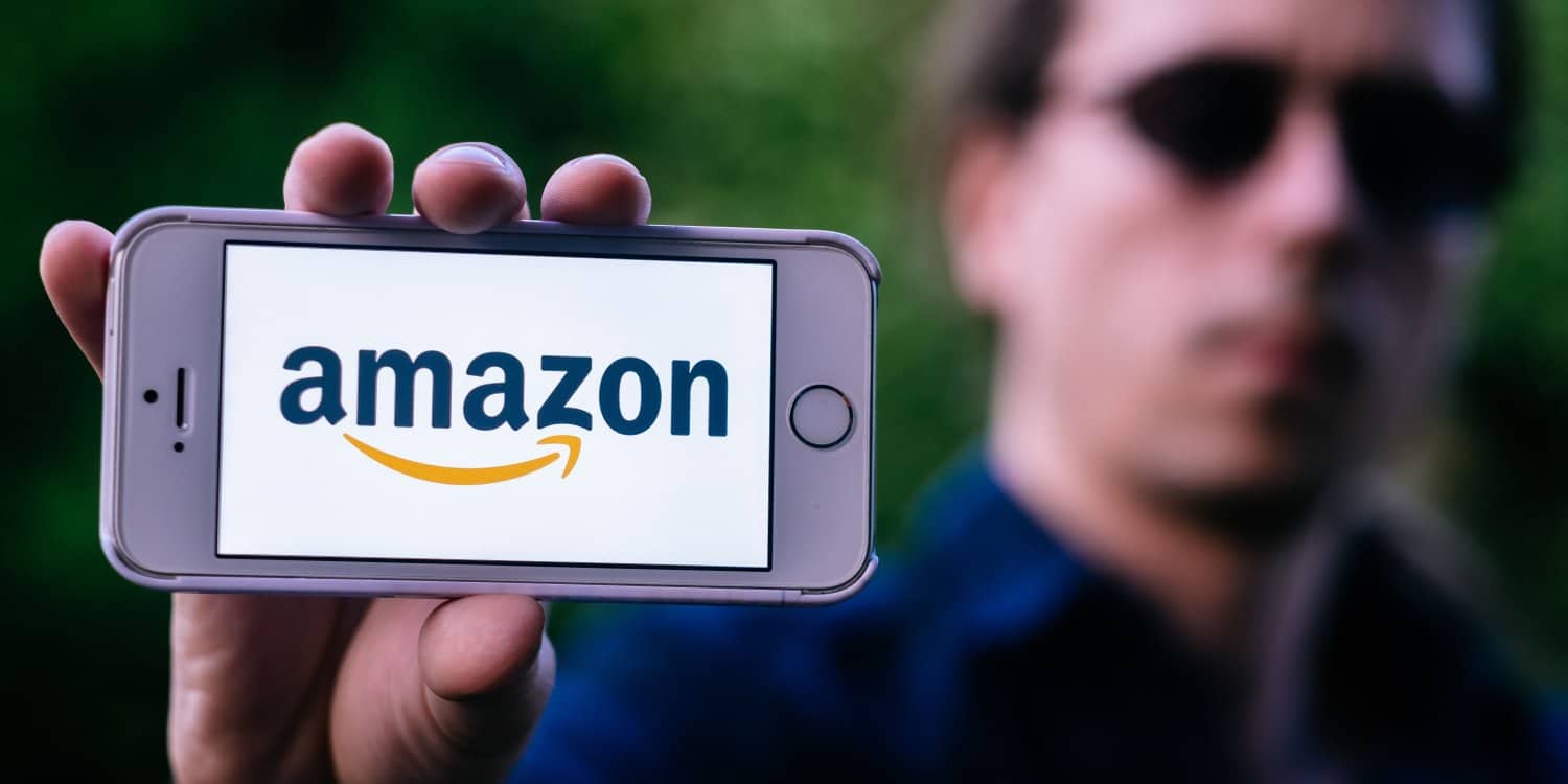 A closeup of a young man wearing dark glasses holding a white iPhone with the AMAZON LOGO on the screen.