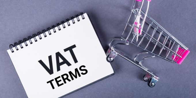 A miniature shopping cart lying next to a white notepad with VAT TERMS written on it in black font.