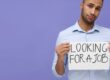 Young unemployed man holding sign with phrase 'Looking For A Job' on violet background, depicting youth unemployment