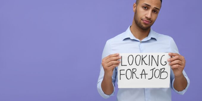 Young unemployed man holding sign with phrase 'Looking For A Job' on violet background, depicting youth unemployment