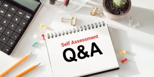 A white notepad with 'Self Assessment Q&A' written on it next to a calculator and other desktop accessories.