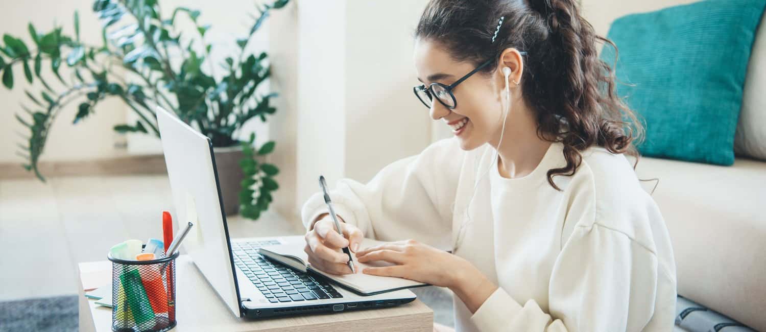 A young girl smiling and sitting in front of a laptop, writing in a notepad, learning about how to open a company as an uner-18