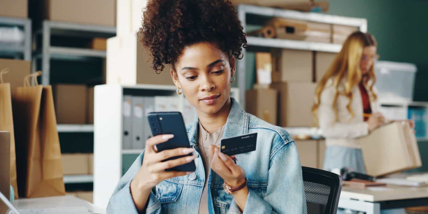 Woman running her business, she's sat at a desk surrounded by boxes being packed up. She's looking at her phone and her credit card, about to pay for something online.