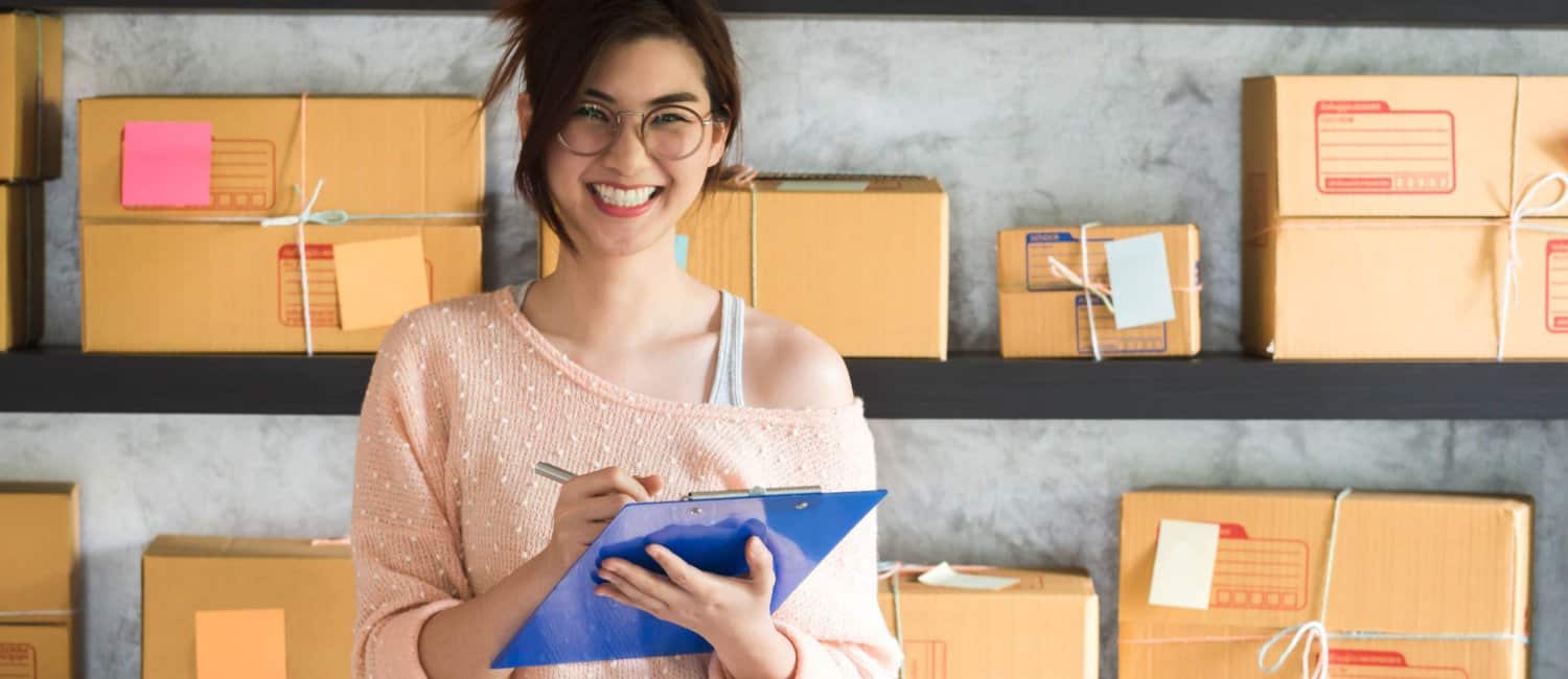 A teenager smiling whilst holding a clipboard and pen, with two shelves of brown boxes behind her, learning about how to open a company as an under-18
