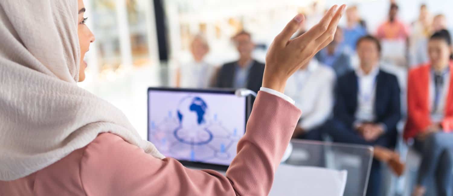 A woman in a pink blazer gestures while presenting International Women's Day to a captivated audience, with an open laptop in front of her.