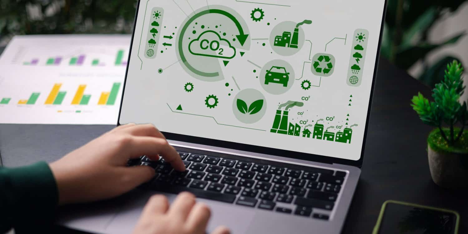 A person's hands on a laptop, with the screen displaying a company's sustainability report. A small plant and desk plant sit next to the laptop and a paper with charts and data is visible behind the laptop