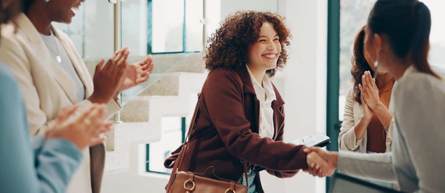 A woman shakes hands with another, while diverse colleagues applaud and celebrate in a bright, modern interior space.