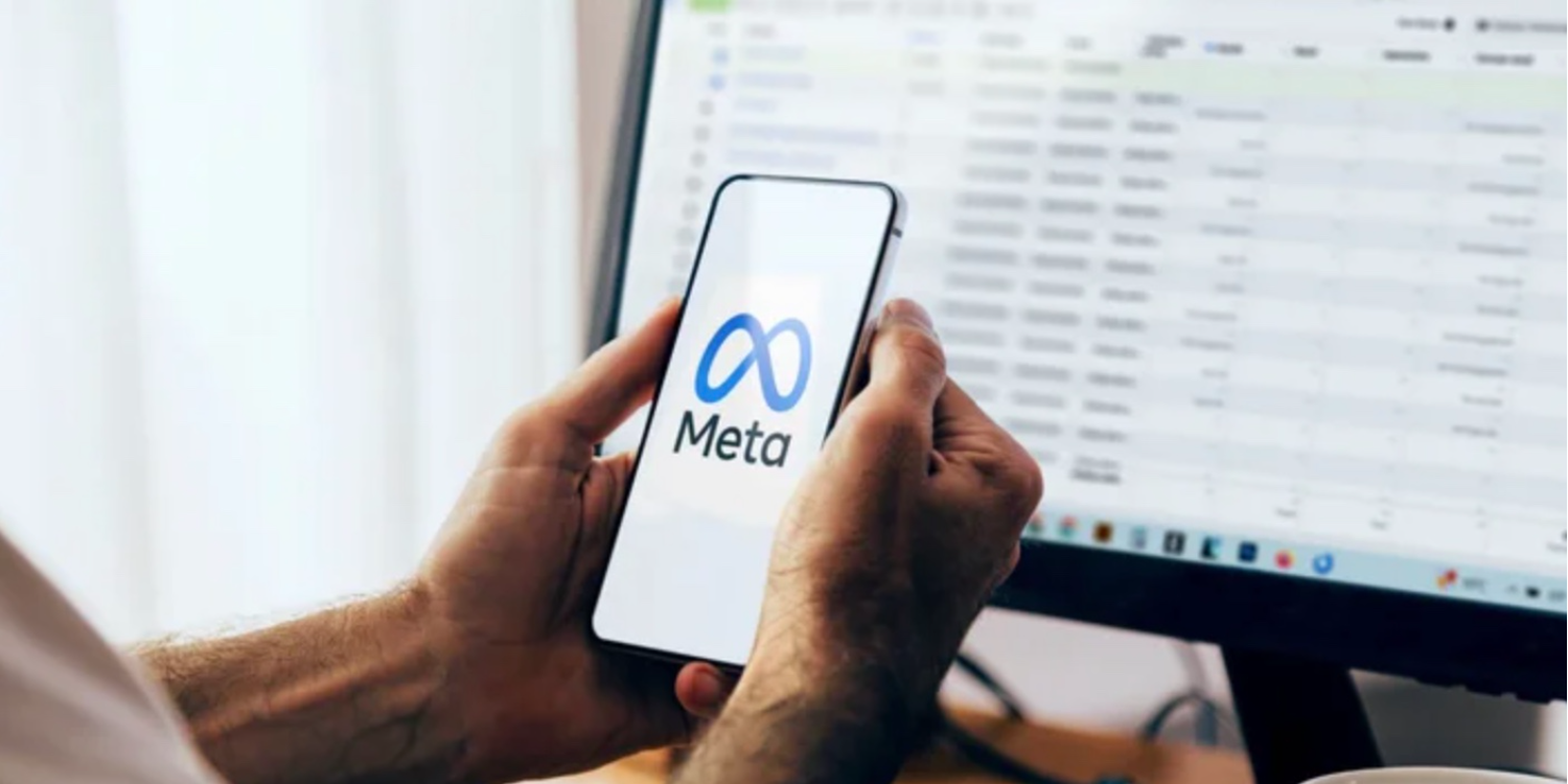Hands holding a smartphone displaying the Facebook Meta logo in front of a data-filled monitor.