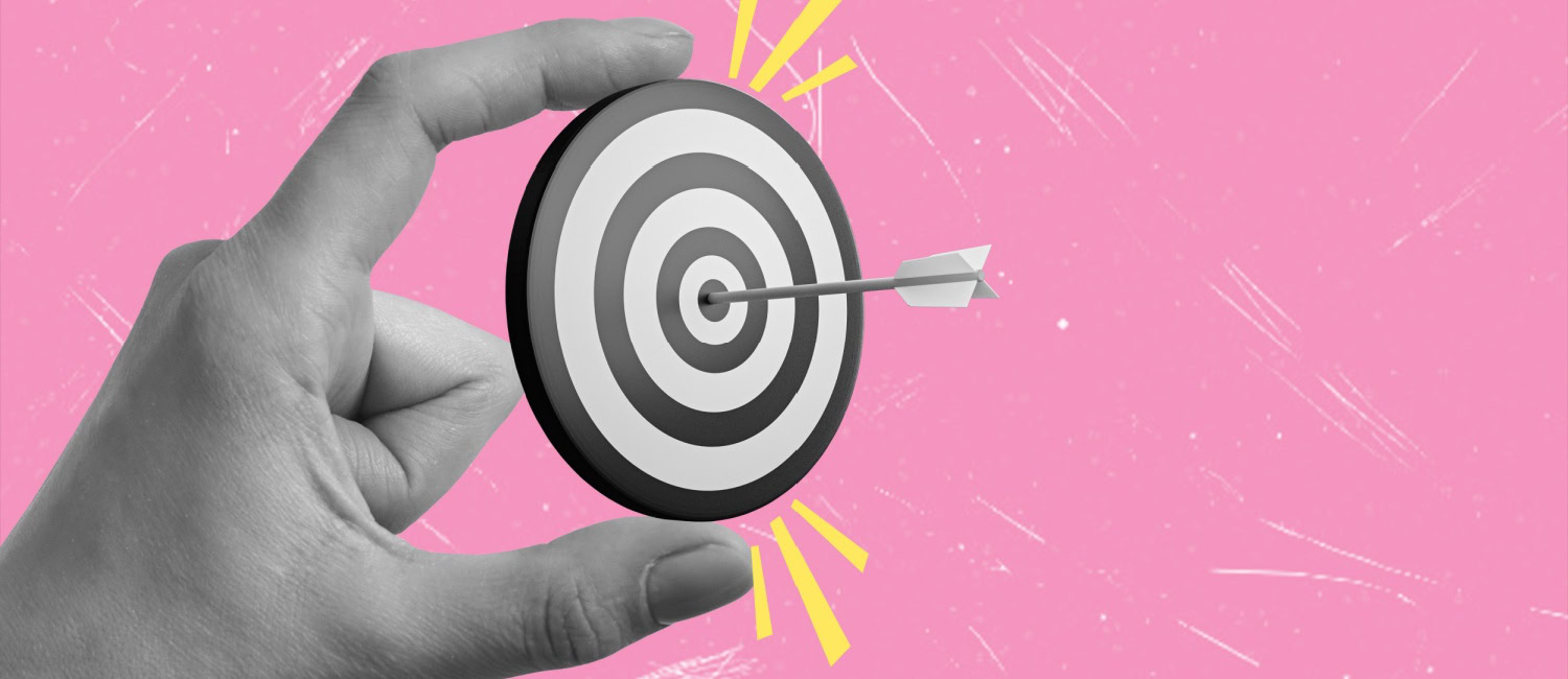 A target held by a hand on a pink background to represent business goals.