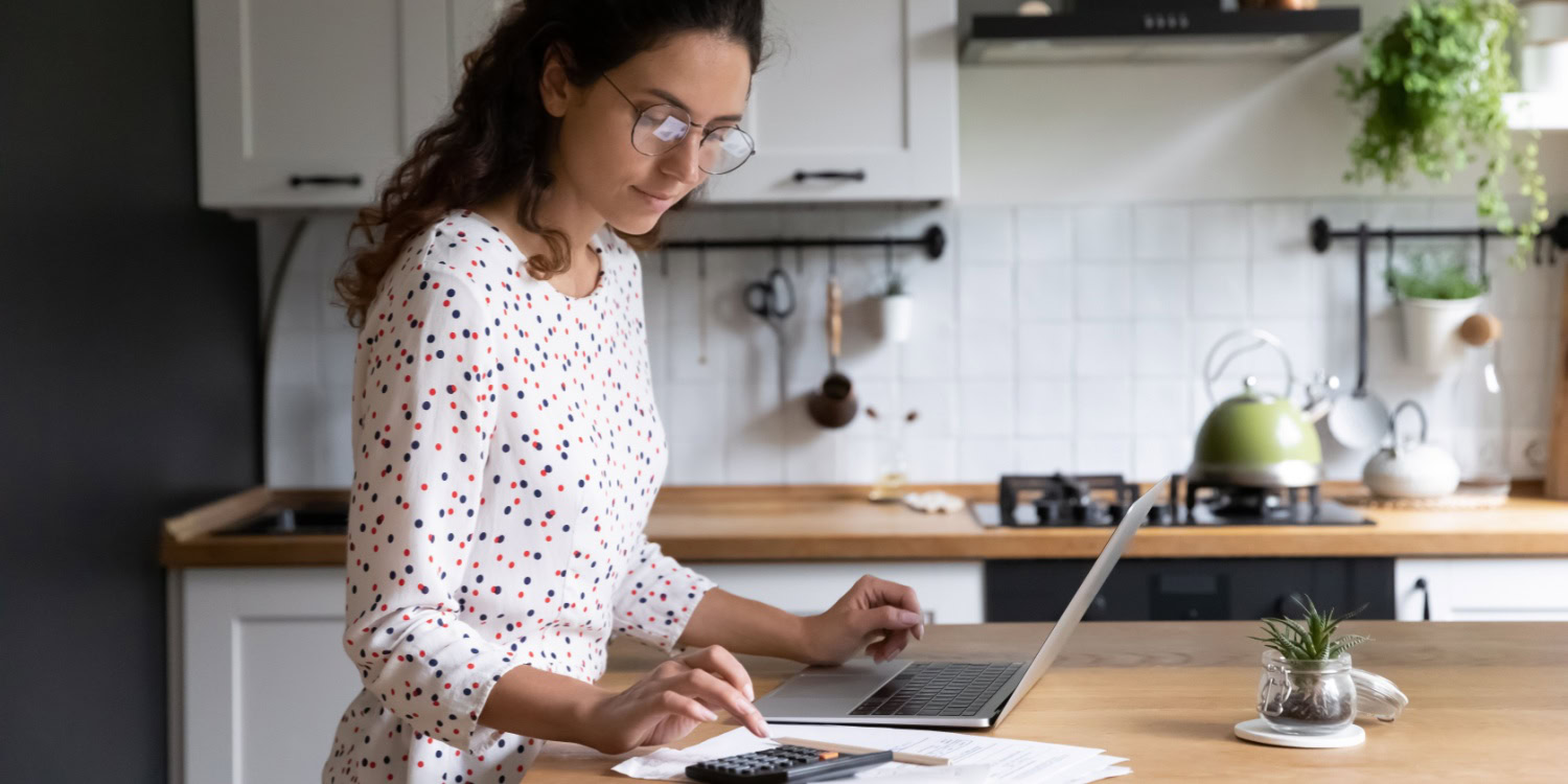Woman working on a laptop in a kitchen, using a calculator and reviewing financial documents, analysing data related to business turnover