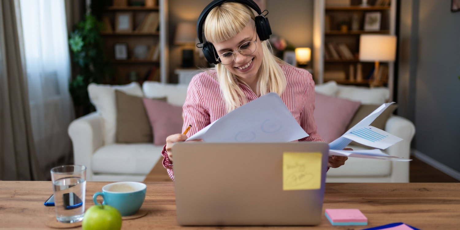 Uni student working on her side hustle from home, wearing headphones and reviewing documents in front of a laptop